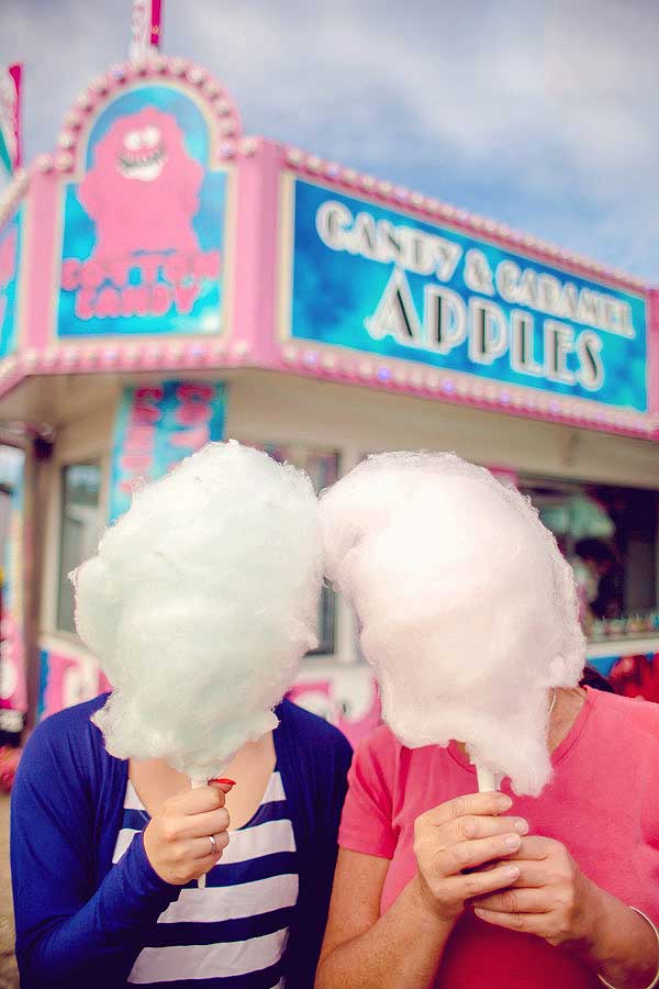 A woman and her daughter both hidden behind cotton candy puffs at the local carnival in front of the food vendor booth in 50's chrome and bright blue and pink colors.